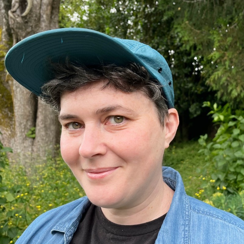 A closeup of Nellie's face, smiling gently, wearing a blue baseball cap, with trees and vegetation behind her